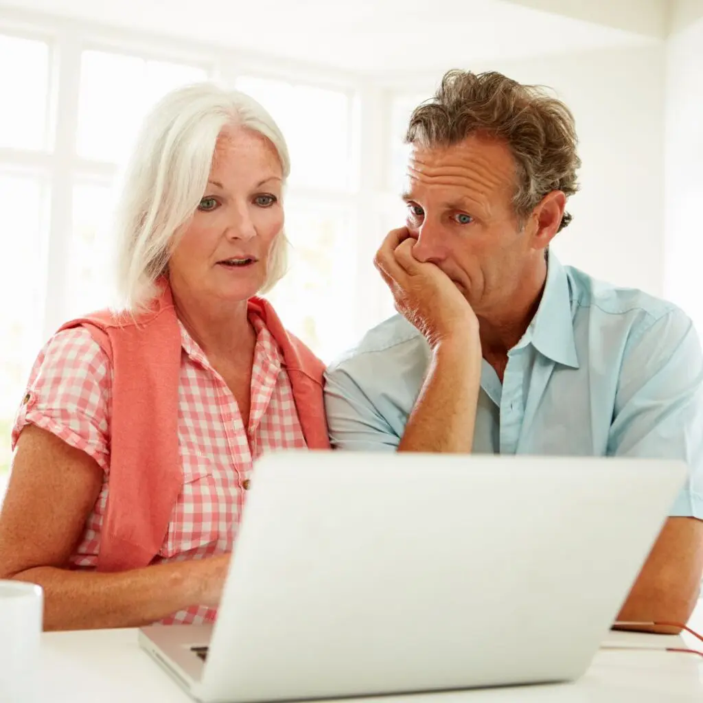 An older couple sit together looking concerned while viewing the screen of a laptop in a bright room after penetrating keratoplasty surgery. The woman wears a pink-checked shirt and the man rests his chin on his hand, both focused on the computer.