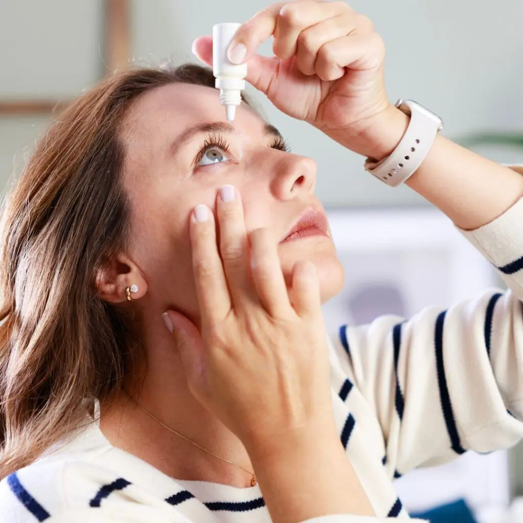 middle aged woman wearing striped top putting eye drops into eye due to one of red eye causes