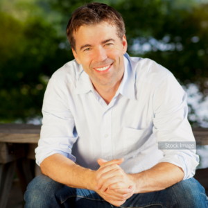 Middle aged man sat outside smiling with pale blue shirt and jeans looking at camera hands clasped