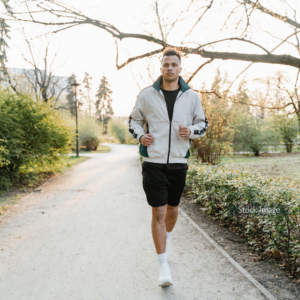 Young man running with black shorts and zip up hoodie in park on path trees and hedges