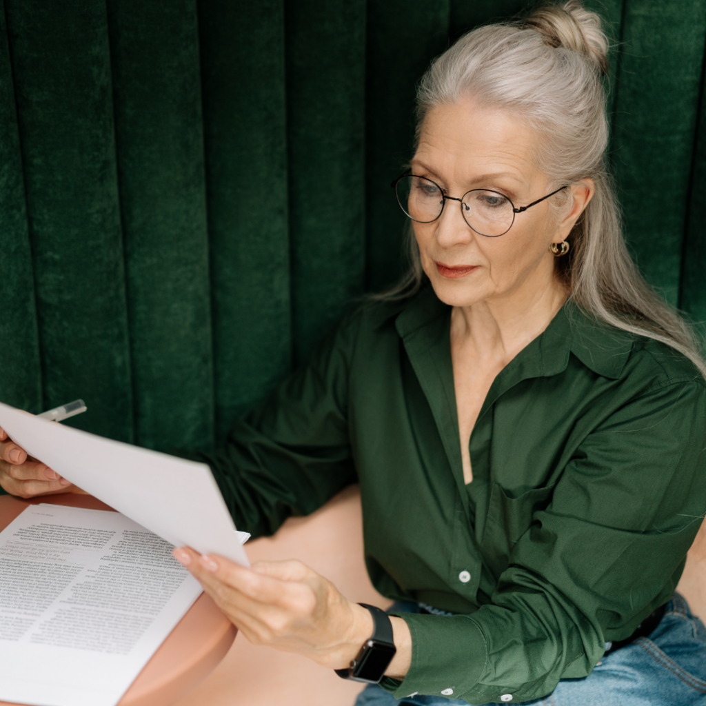 Older lady with grey hair and reading glasses wearing evergreen shirt and reading sheet of paper with pen for blog post on presbyopia and the need for reading glasses