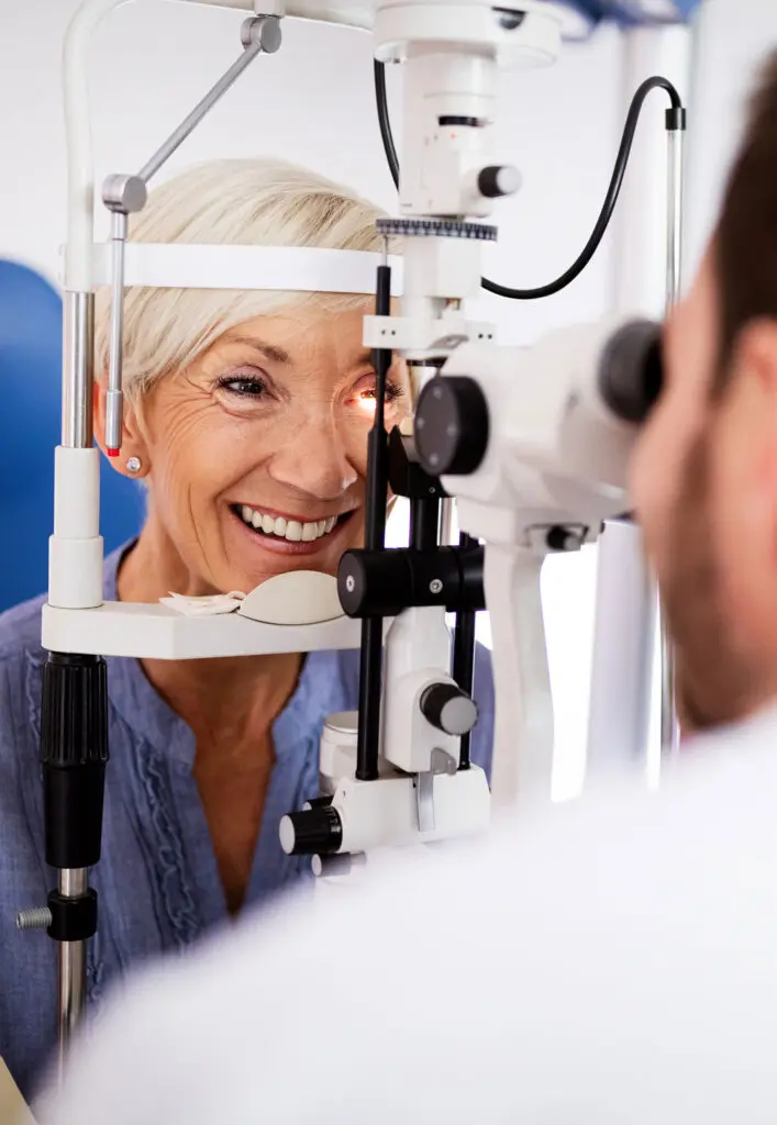 An older woman smiles while having her eyes examined by an optometrist using a slit lamp in a clinic. The optometrist’s back is to the camera.