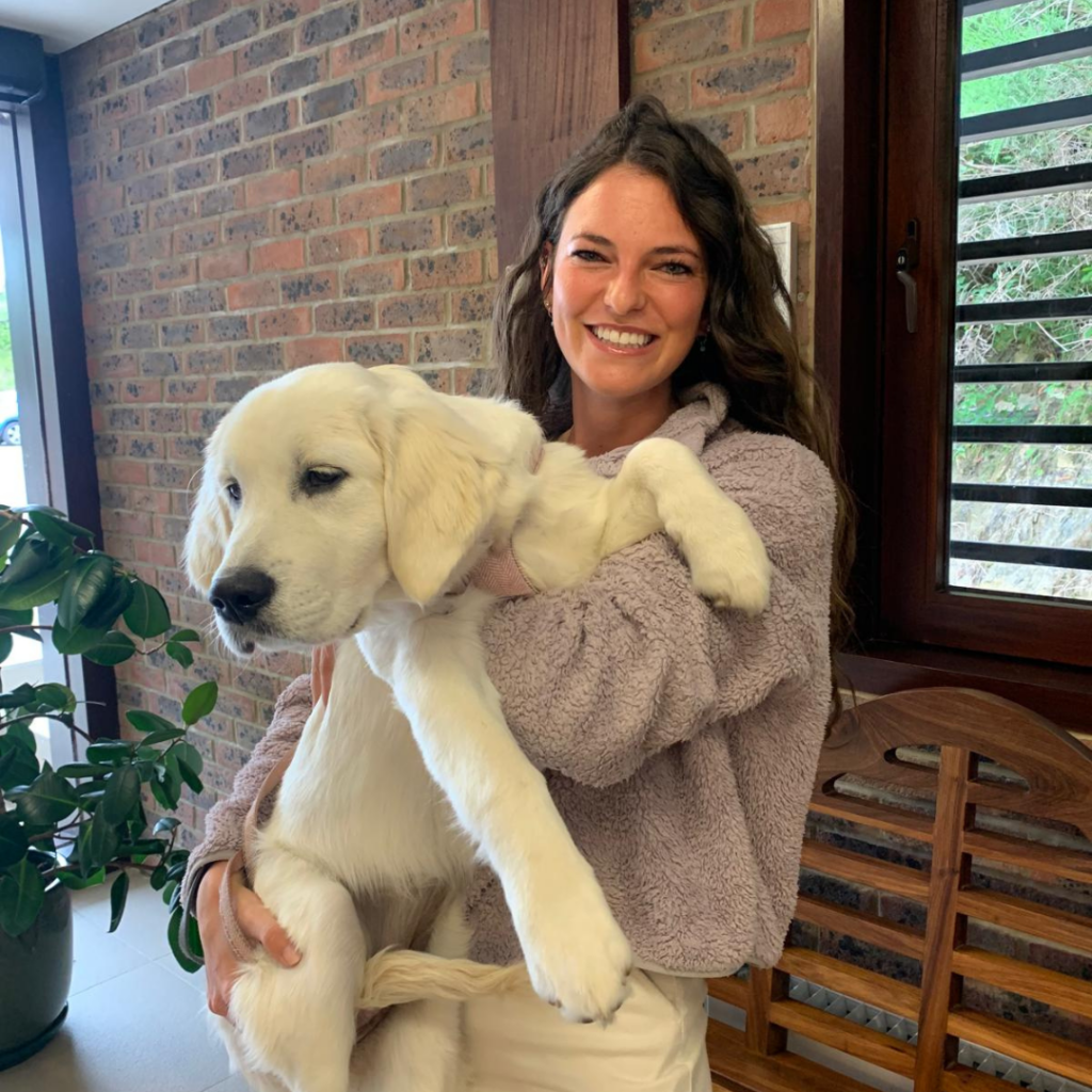 A smiling woman with long dark hair holds a large, fluffy white dog indoors at a Patient Event near a window and a brick wall, with a green potted plant beside her.