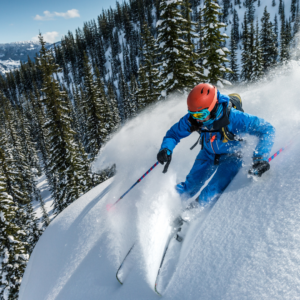 Skier in mountains with trees in the background wearing orange helmet and blue ski suit on the move