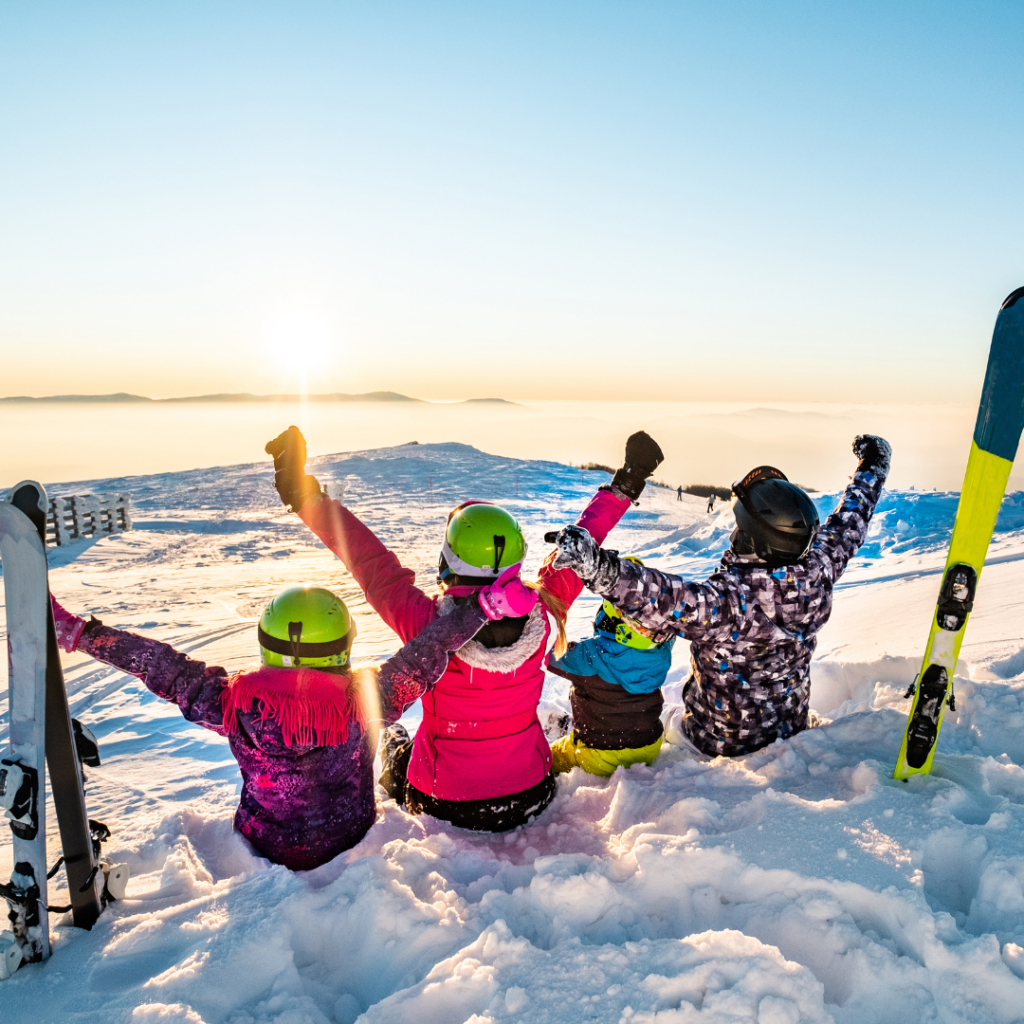 Family in the mountains whilst skiing sitting down looking a the sunset view with arms in the air happy