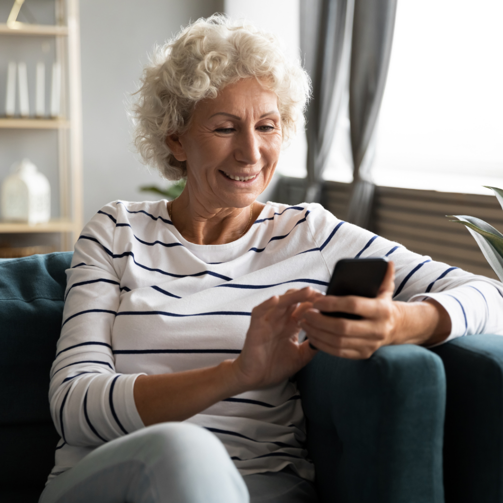 Older lady with grey hair looking at phone on sofa smiling after 2RT laser eye treatment
