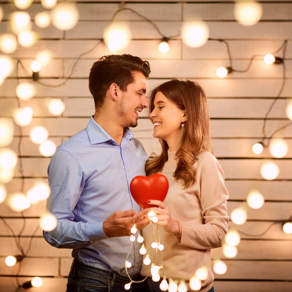 Young couple man and woman holding and smiling at each other surrounded by fairy lights holding a red heart