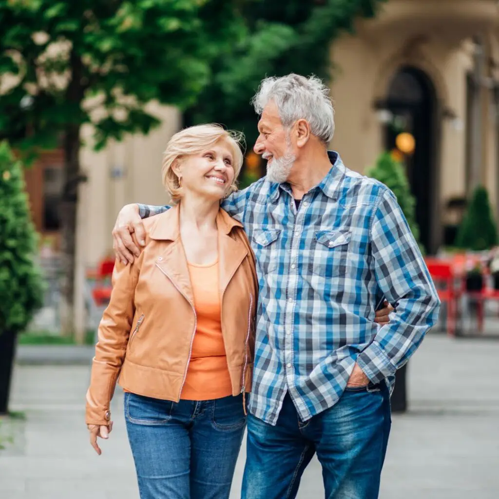 An older couple, smiling at each other after laser cataract surgery, enjoy a walk outside. The woman wears a tan jacket and orange shirt, while the man sports a blue checked shirt. Trees and outdoor seating are in the background.