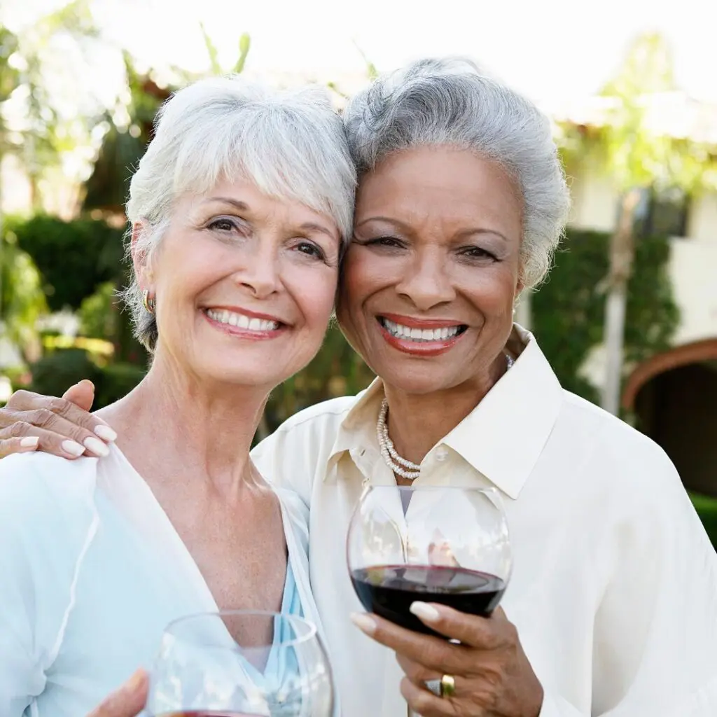 Two older women smiling and embracing outdoors, each holding a glass of red wine, enjoying life after laser cataract surgery, with greenery and a building in the background.