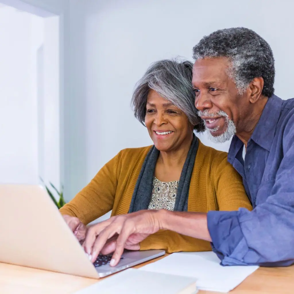 An older couple smiles whilst sitting together at a table, looking at a laptop screen. The woman, newly confident after laser cataract surgery, types on the keyboard as the man leans in, both appearing happy and engaged.