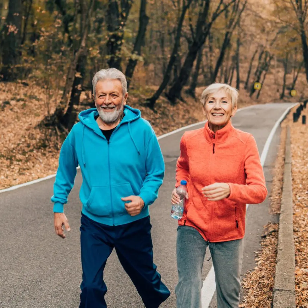 An older man and woman smile whilst walking on a paved path through a forest after refractive cataract surgery at Centre for Sight. The man wears a blue hoodie, and the woman, in an orange jacket holding a water bottle, enjoys vibrant autumn leaves underfoot.