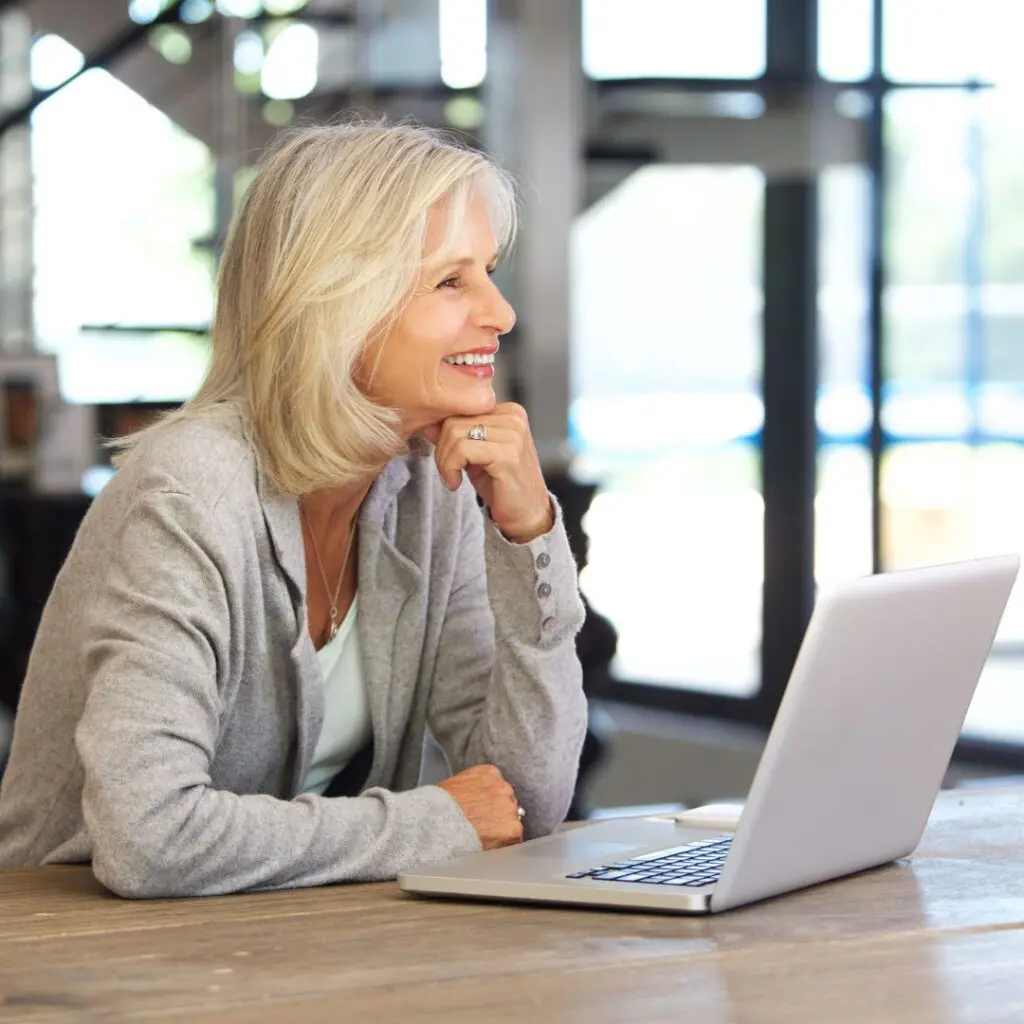 A smiling older woman with grey hair, who recently had refractive cataract surgery at Centre for Sight, sits at a table, looking at a laptop in a bright, modern room. She rests her chin on her hand and appears engaged with what’s on the screen.