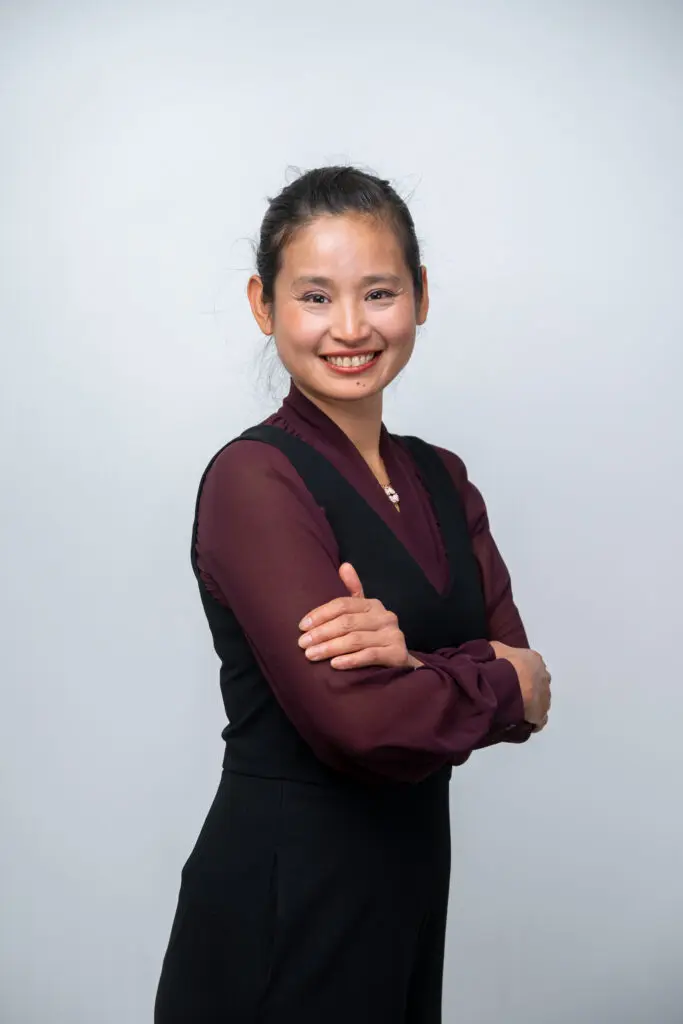 A woman with dark hair tied back smiles at the camera, standing with arms folded. She wears a maroon long-sleeved top under a black sleeveless dress, against a light grey background.