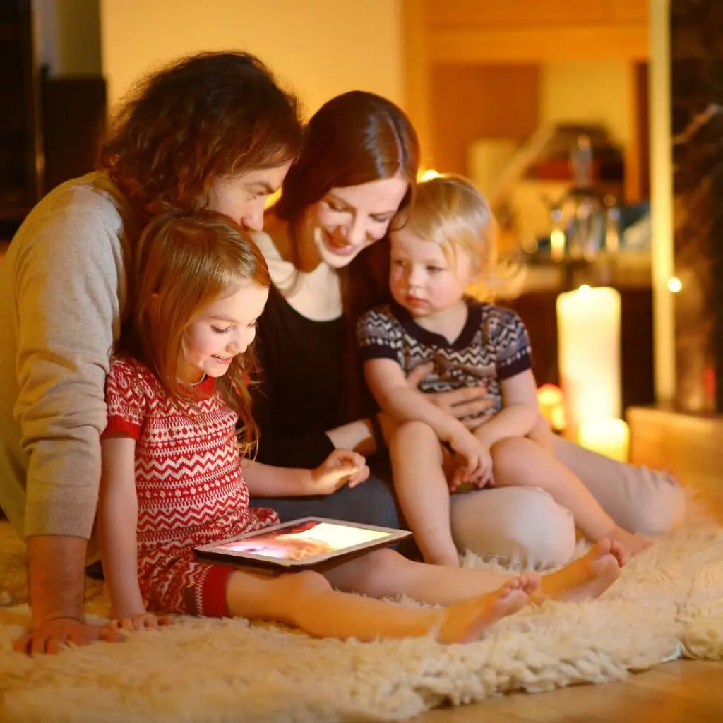 A family of four sits together on a cosy rug, illuminated by warm light. The young girl holds a tablet, whilst her parents and younger sibling watch, smiling and enjoying time together indoors.