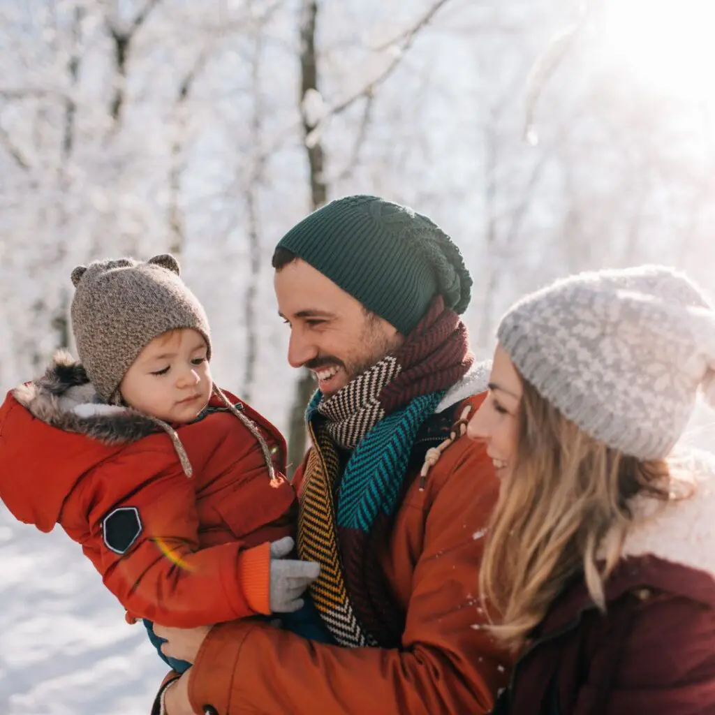A smiling couple in winter hats and coats hold a young child dressed warmly, standing outdoors in a snowy forest with sunlight shining through the trees.