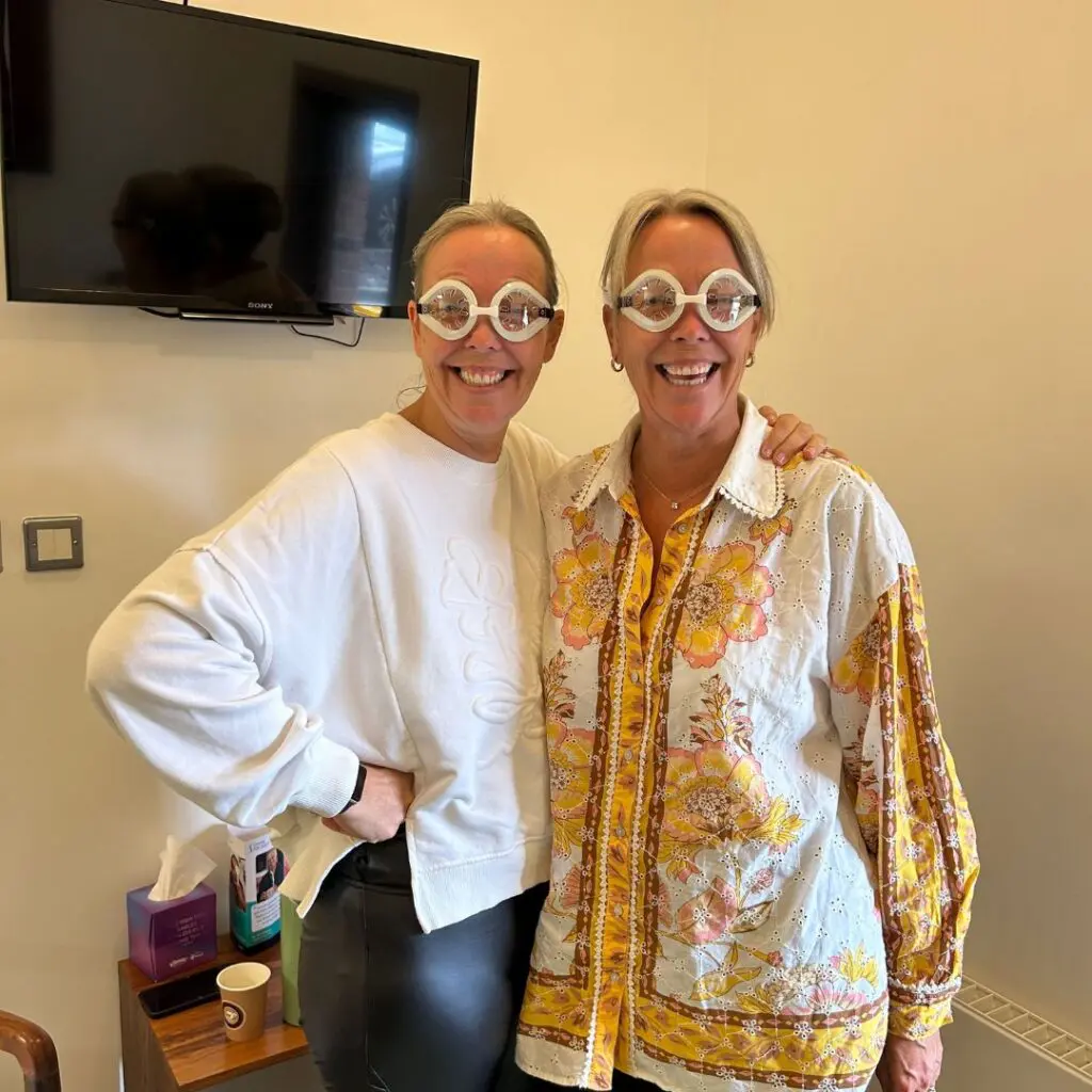 Two smiling women stand indoors wearing fun oversized, twin circular glasses. One wears a white sweatshirt and black trousers, while the other wears a floral shirt. A telly and tissue box can be seen in the background.