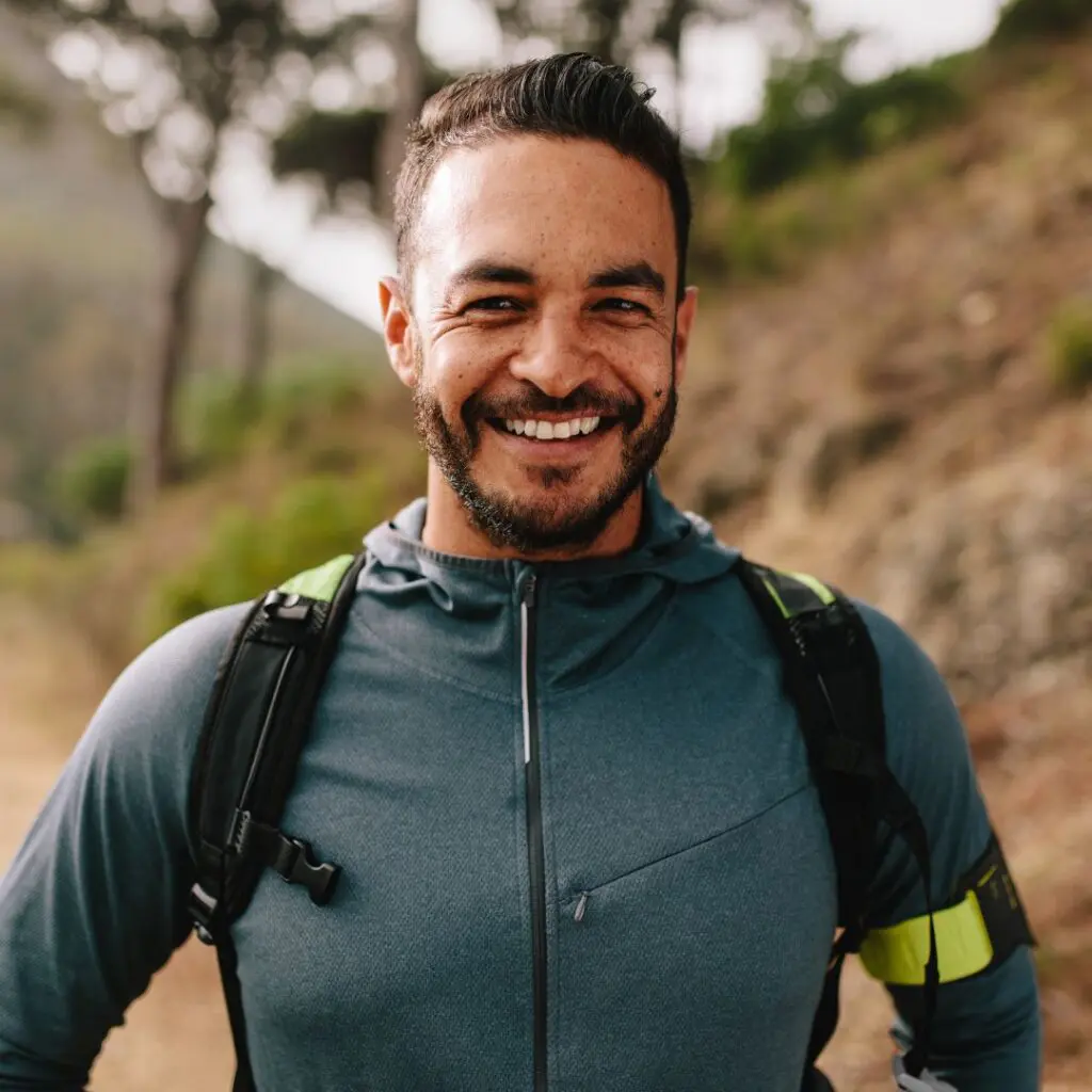 A man with short dark hair and a beard smiles whilst wearing a blue jacket and rucksack, standing on a hiking trail with blurred trees and greenery in the background after LEX laser eye surgery at Centre for Sight.