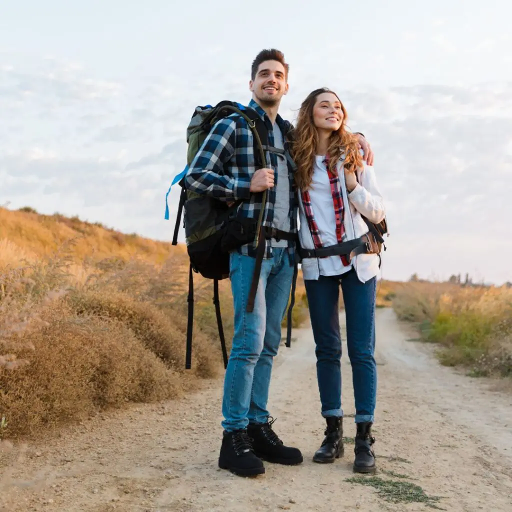 A smiling young couple in casual hiking clothes and rucksacks stands on a dirt path in a grassy open field, looking off into the distance under a partly cloudy sky after LEX laser eye surgery at Centre for Sight.