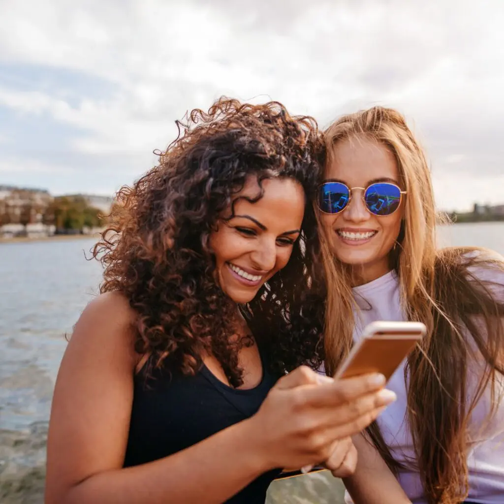 Two women smiling and looking at a mobile phone together outdoors by the water. One has curly hair, the other has straight hair and is wearing blue reflective sunglasses. It’s a bright, sunny day after LEX laser eye surgery at Centre for Sight.