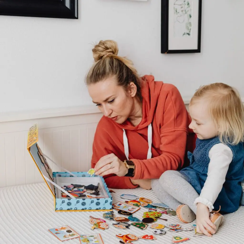 A woman in an orange hoodie and a young child sit on a bed, playing with colourful cardboard pieces from a game or puzzle. The woman is focused on the pieces, while the child watches intently after LEX laser eye surgery at Centre for Sight.