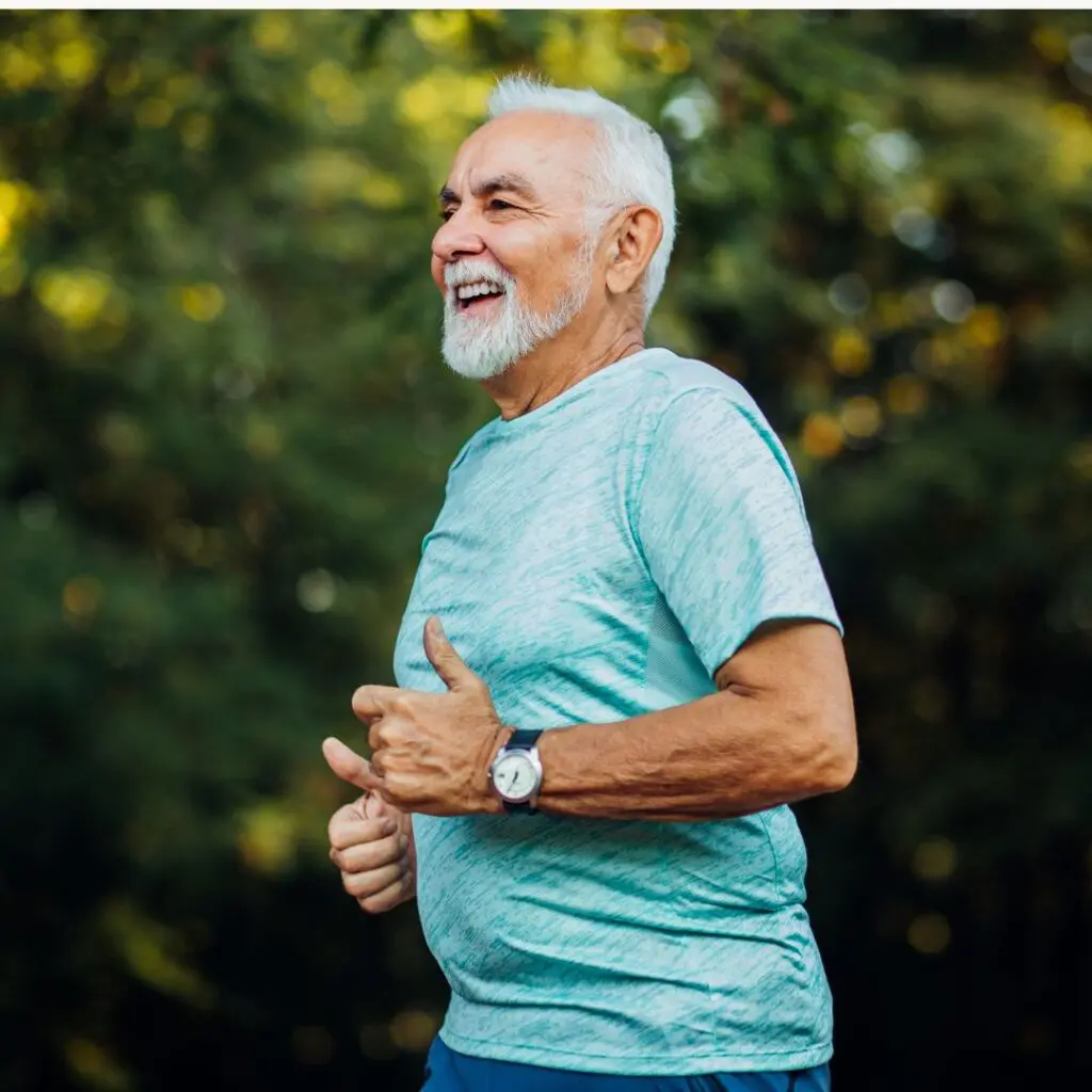 An older man with grey hair and a beard is jogging outdoors, smiling and wearing a light blue sports shirt and a watch, with green trees blurred in the background.