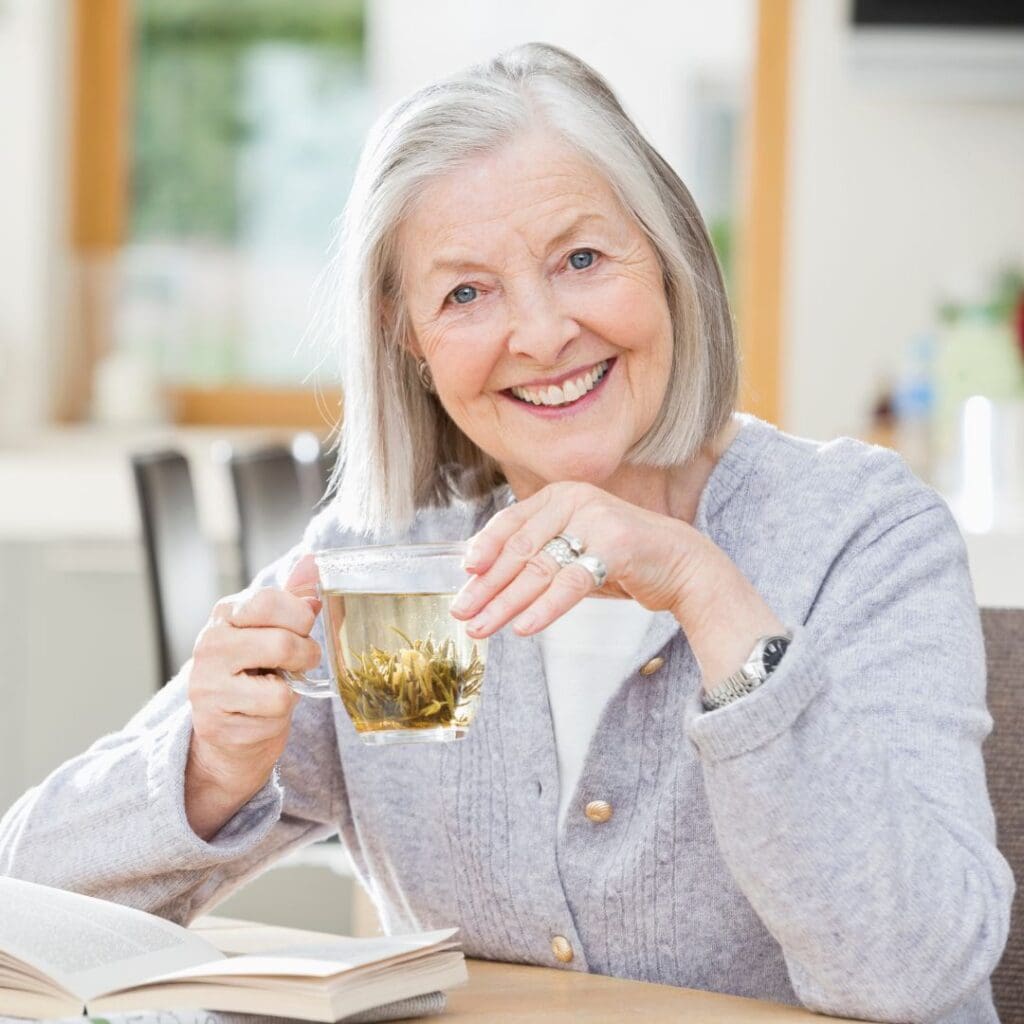 Centre for Sight older lady with grey hair wearing grey jumper sipping herbal tea and smiling as she reads her book looking straight at camera stock image