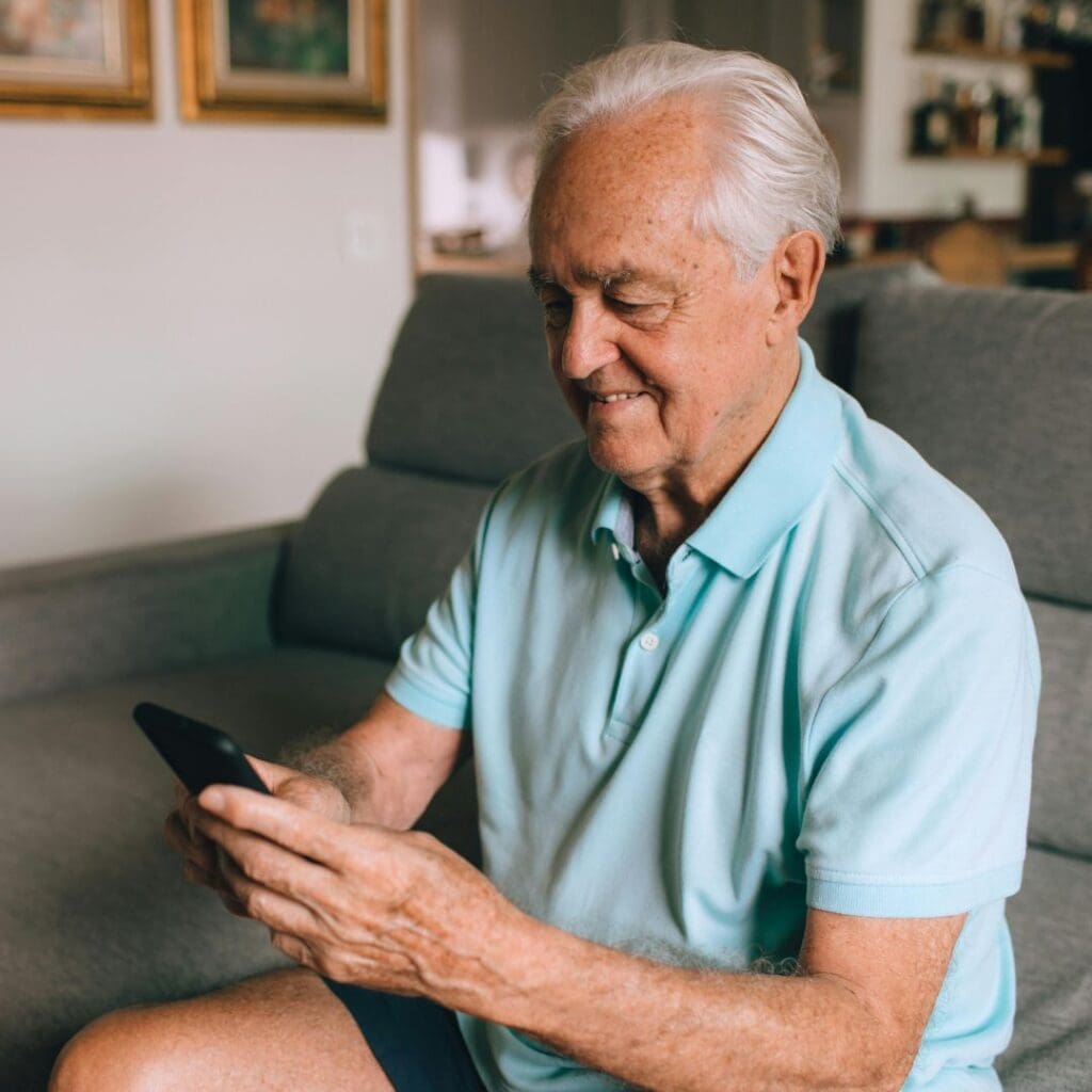 Centre for Sight older man sat on sofa in house wearing blue t shirt looking at smartphone smiling stock image grey hair balding