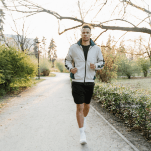 Young man running with black shorts and zip up hoodie in park on path trees and hedges