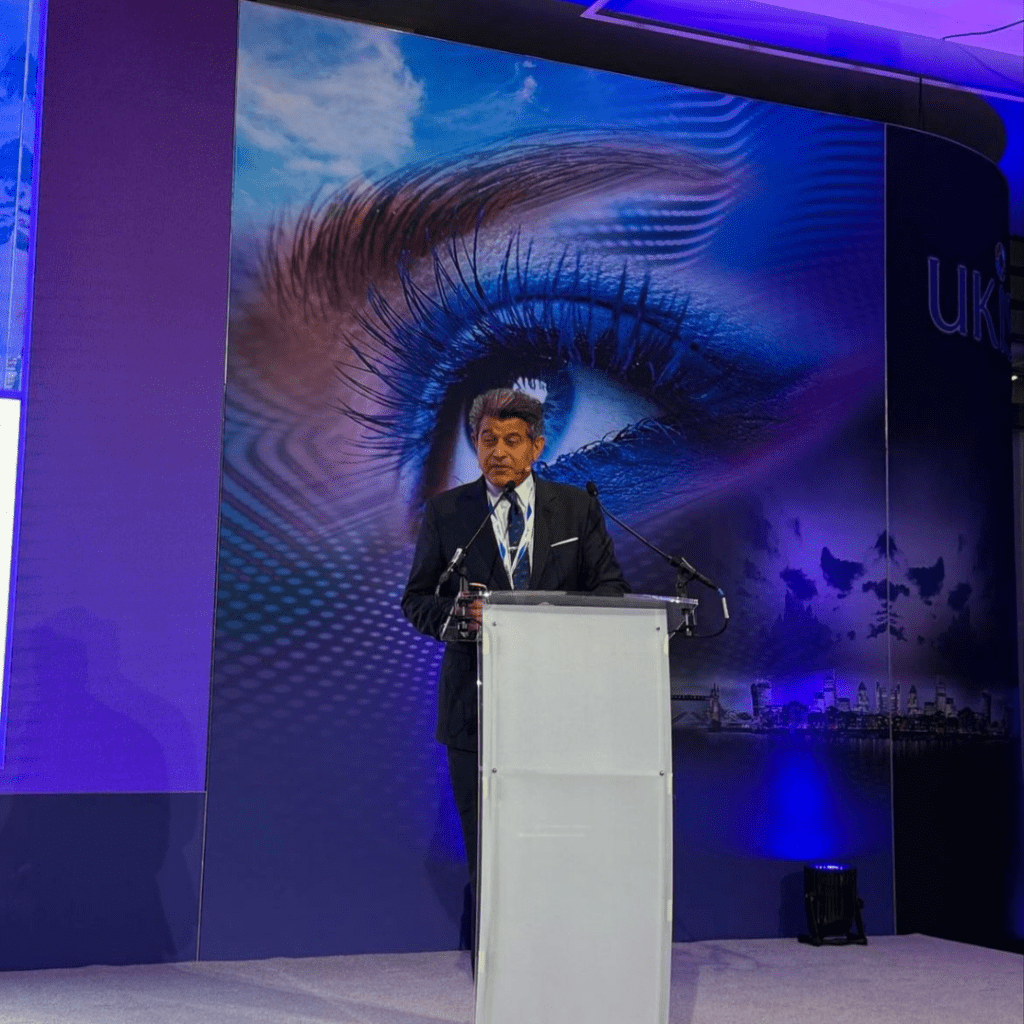 Man on stage speaking at a podium speaking into a microphone with eye in the background at event