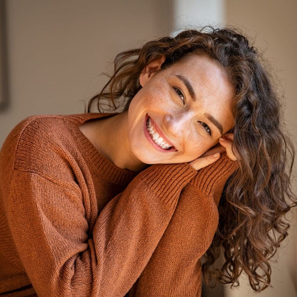 Centre for Sight young woman with brown jumper and brunette curly hair smiling with hands on her check
