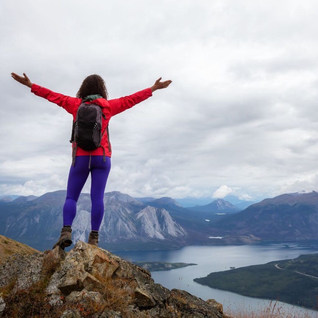 Centre for Sight hiking woman in red jacket and backpack standing on cliffside arms stretched out freedom with valley below river and scenic views