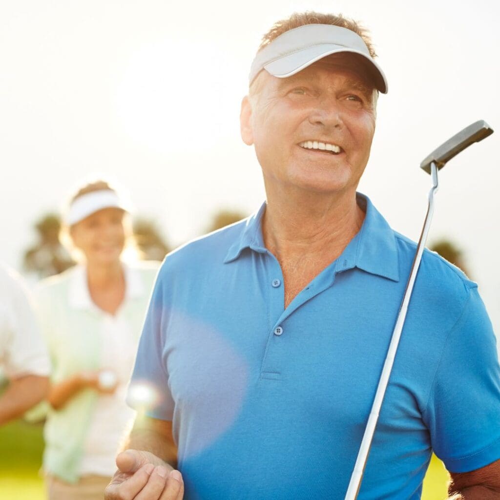 Centre for Sight older man with golf hat on on the course with friends holding putter in blue golf top smiling and looking out over course behind camera