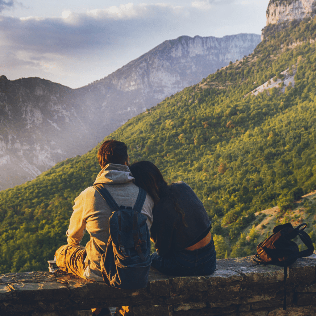 Centre for Sight young couple in coats and backpack brunette hair looking out over view of trees sitting on wall cuddling backs to camera