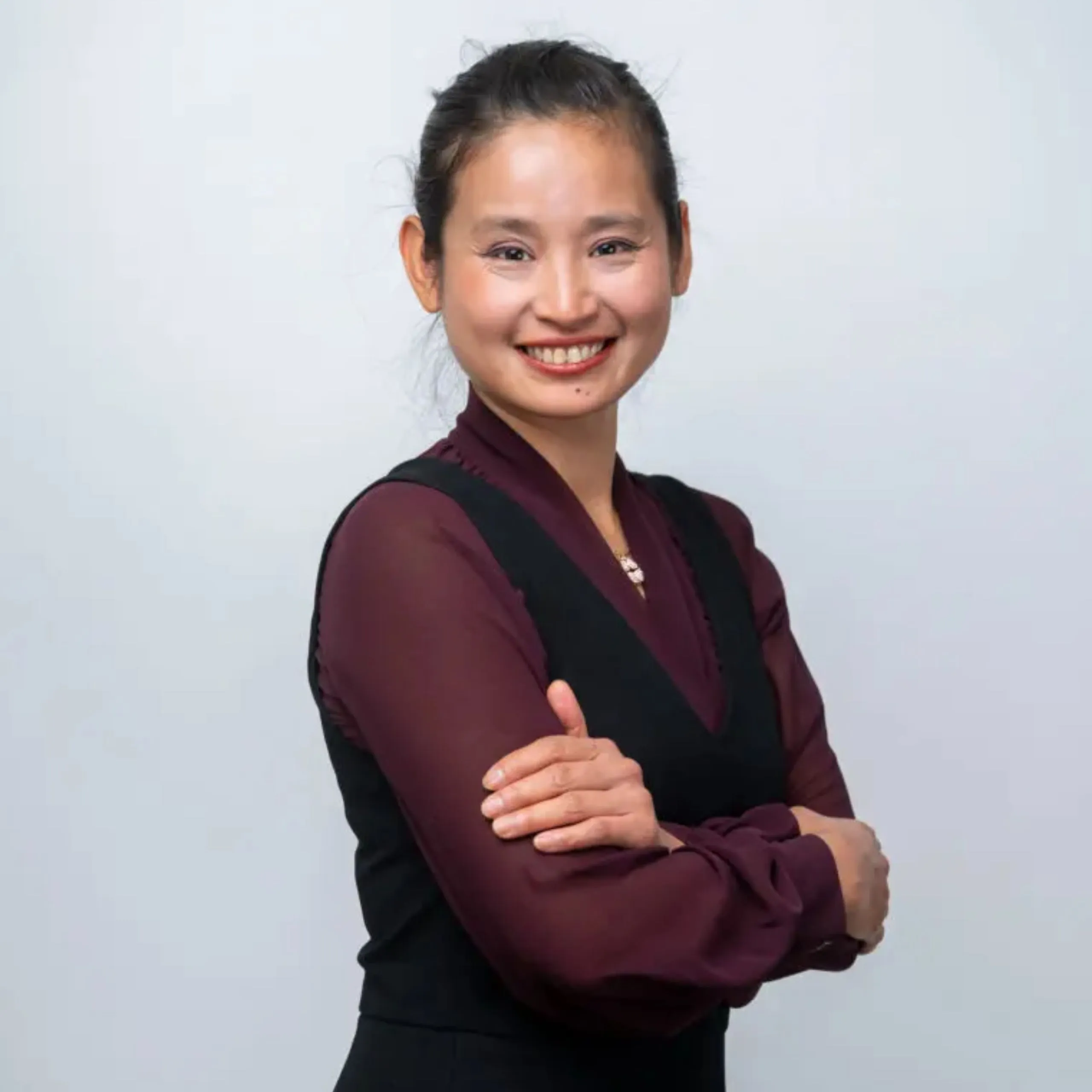 A woman with dark hair tied back, wearing a burgundy blouse and black waistcoat, stands smiling with her arms crossed in front of a plain light background.