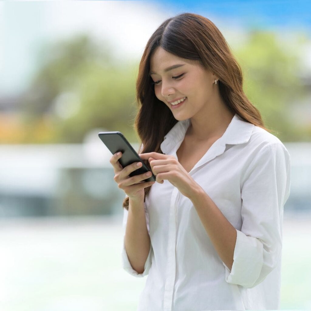 Is Laser Eye Surgery Painful? 10 Centre for Sight young girl with dark brunette hair in white shirt looking at mobile phone researching and smiling|Centre for Sight young woman with brown jumper and brunette curly hair smiling with hands on her check